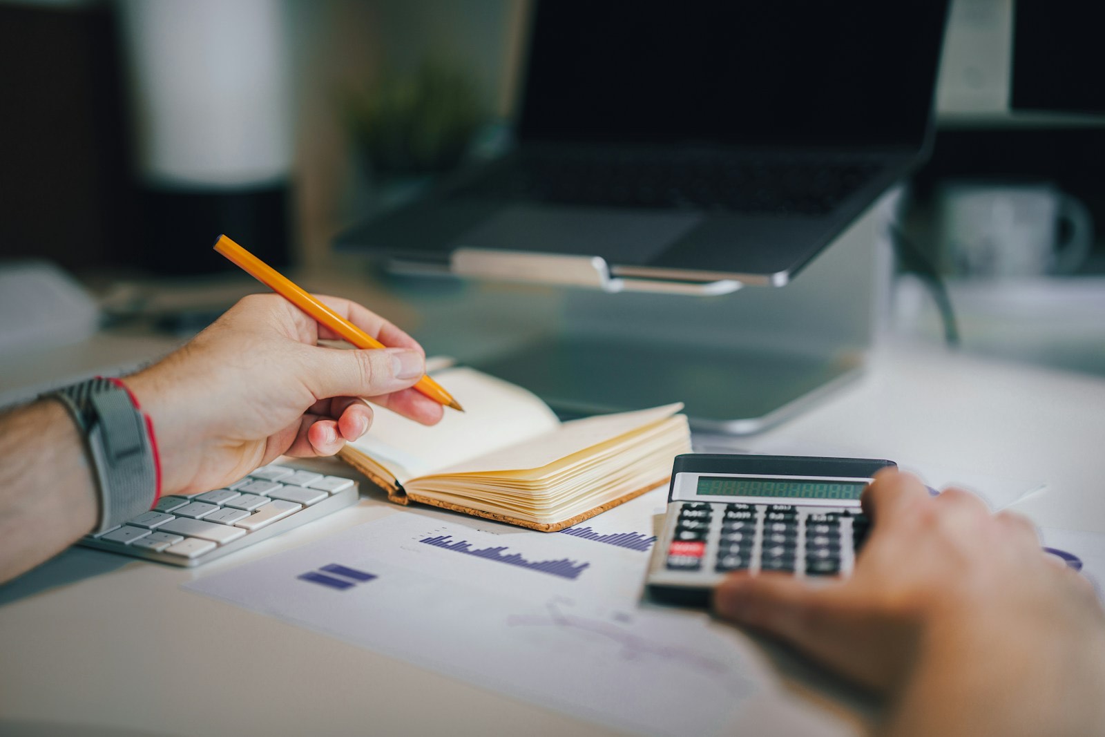 Manos con un lápiz y una calculadora sobre una libreta y hojas con gráficas