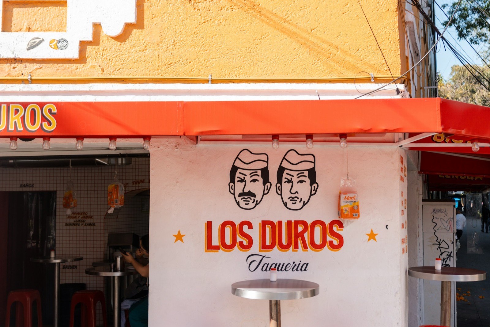 Esquina de una taquería con toldo rojo, mesas altas y bancos naranjas en la banqueta